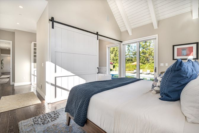 Sunlit modern bedroom with vaulted white ceiling, sliding barn door, navy throw and pillows on a crisp bed, and glass doors opening to a backyard pool view.