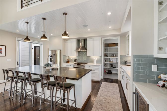 Bright open-concept modern kitchen with a large dark island and five metal bar stools, white shaker cabinets, stainless steel appliances, glass subway-tile backsplash, hanging pendant lights, dark hardwood floors and a walk-in pantry.