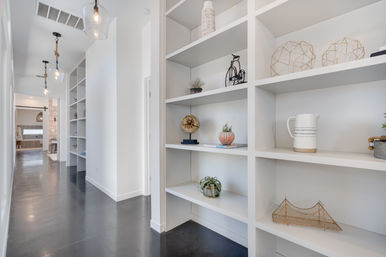 Sleek modern hallway with white built-in shelves displaying plants, pottery and geometric decor, pendant lights and polished dark concrete floor leading to an open living area
