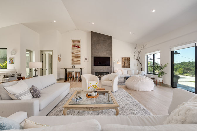 Bright sunlit modern open-concept living room with vaulted ceiling, neutral sofas and armchairs around a wooden coffee table, gray feature fireplace wall, large sliding glass doors and windows overlooking greenery, plush rug, pouf beanbag and minimal decor.