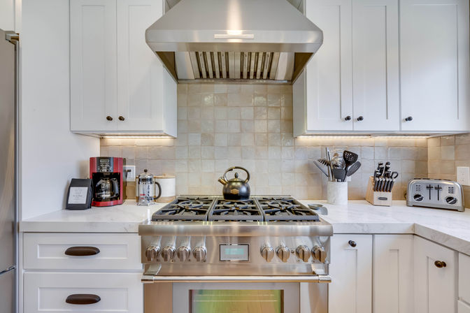 Bright modern home kitchen with stainless steel gas range and vent hood, black kettle on the stove, white shaker cabinets, tiled backsplash and countertop appliances like a coffee maker, French press, toaster and knife block.