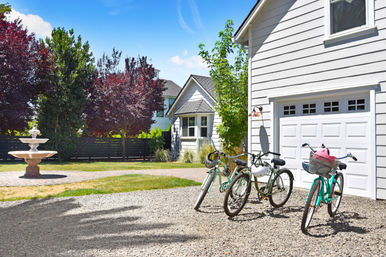 Three bicycles parked on a gravel driveway beside a white garage of a suburban house, with a stone fountain, landscaped lawn and leafy trees under a bright blue sky