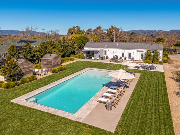Rectangular swimming pool in a sunny backyard oasis with lined sun loungers and umbrellas, manicured green lawn, modern white pool house and wooden cabanas