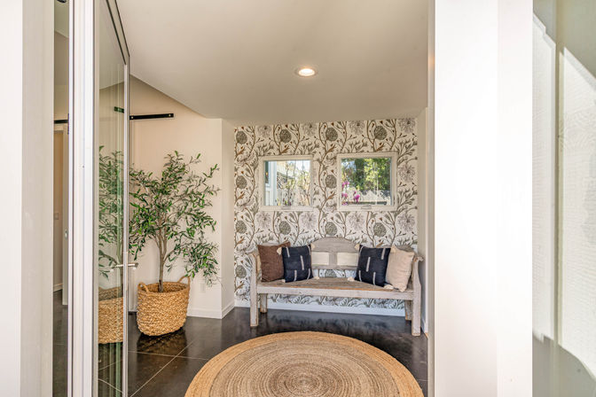 Cozy entryway nook with distressed wooden bench, neutral and navy throw pillows, botanical wallpaper behind two small windows, round woven jute rug on dark polished floor, and a potted leafy plant in a wicker basket.