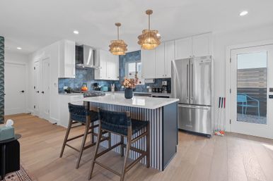 Bright modern coastal kitchen with blue hex tile backsplash, white cabinets, large quartz island with navy fluted base, two woven navy bar stools, rattan pendant lights, stainless steel fridge and light hardwood floors
