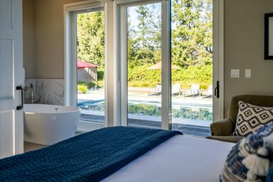 Modern master bedroom with freestanding bathtub beside sliding glass doors that open to a sunlit backyard pool, poolside loungers and umbrellas, navy bed throw and a cozy armchair with patterned pillow.