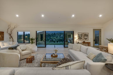 Modern open-plan living room with neutral sofas and armchairs, glass coffee table on hardwood floors, large sliding glass doors opening to a lush hillside view and patio under warm recessed lighting.