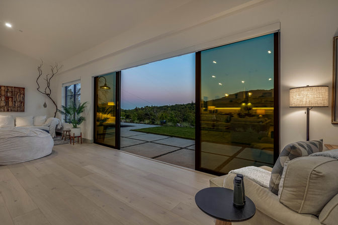 Modern open-plan living room with pale wood floors, plush seating and round side table, potted plant and floor lamp, large sliding glass doors opening to a patio and rolling hillside at twilight.