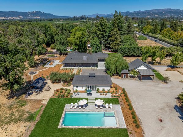 Aerial view of a sunny luxury country estate with a rectangular sparkling pool, poolside loungers and umbrellas, main house and guest cottage, landscaped patios, tree-lined driveway and rolling hills in the distance.