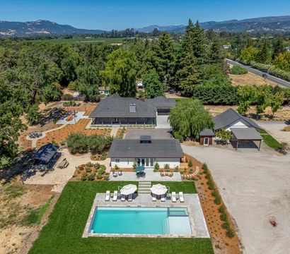 Aerial view of a sunny luxury country estate with a rectangular sparkling pool, poolside loungers and umbrellas, main house and guest cottage, landscaped patios, tree-lined driveway and rolling hills in the distance.