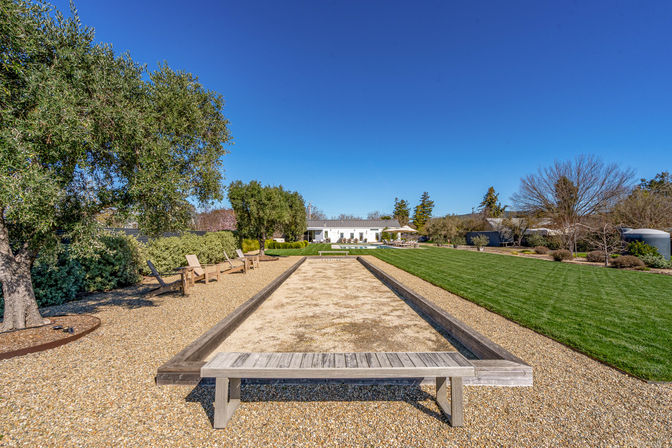 Sun-drenched backyard bocce court with a wooden bench, gravel seating area and Adirondack chairs under olive trees, manicured green lawn and modern white house under a clear blue sky.