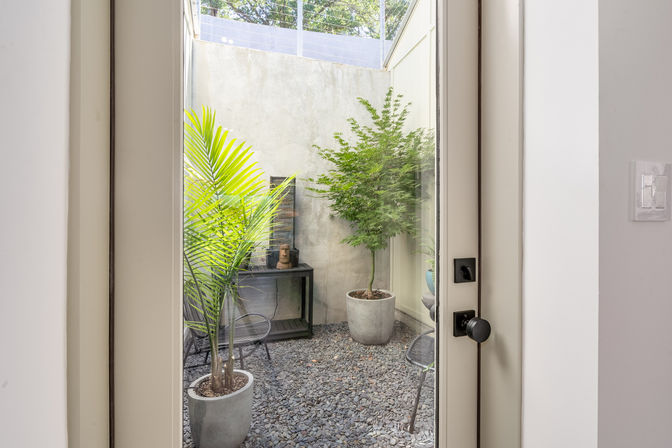 View through a glass door into a cozy urban courtyard with pebble floor, concrete walls, potted palm and small leafy tree, black metal console table and patio chairs — modern minimalist small patio.
