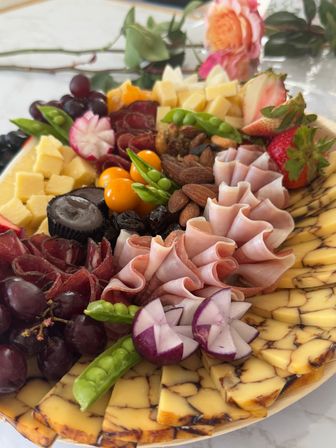 Gourmet charcuterie board with marbled cheese slices, folded ham and cured meats, cubed cheddar, grapes, strawberries, nuts, pea pods and carved radishes on a marble tabletop with roses.