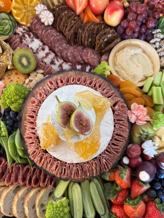 Overhead view of a vibrant charcuterie grazing board centered on a wheel of brie topped with halved figs and honeycomb, surrounded by folded salami, seeded crackers, grapes, strawberries, kiwi, cucumber spears, apricots, hummus and sliced baguette.