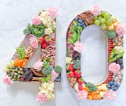 Festive '40' shaped charcuterie board on white marble, packed with strawberries, blueberries, grapes, blackberries, dried apricot, assorted cheeses, crackers, nuts, snap peas, broccoli florets and pink carnations — colorful 40th birthday grazing board.