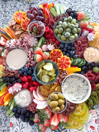 Vibrant charcuterie grazing board with bowls of green olives and pickles, creamy dips, cured meats, strawberries, grapes, blueberries, raspberries, blackberries, sliced cucumbers, radishes, bell peppers and assorted nuts on a speckled granite surface