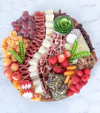 Overhead view of a vibrant round charcuterie board on marble with folded cured meats, assorted cheeses, grapes, strawberries, raspberries, cherries, dried apricots, snap peas, cucumber rosette, nuts, chocolate-pistachio clusters and crackers — a gourmet cheese platter for entertaining.
