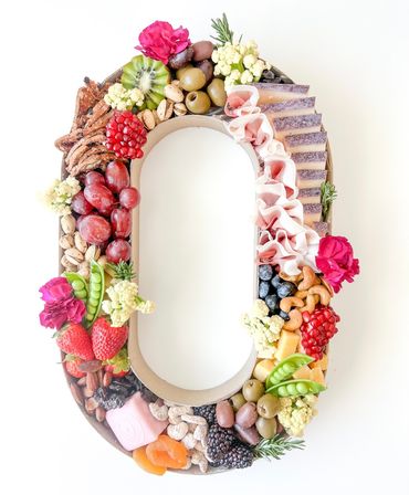 Oval wreath-style charcuterie grazing board on white background with cheeses, folded cured meats, grapes, strawberries, olives, nuts, berries, snap peas and edible flowers — colorful party platter for entertaining.