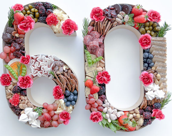 60-shaped charcuterie grazing board arranged with strawberries, grapes, blackberries, blueberries, assorted cheeses, cured meats, crackers, nuts, chocolates, meringues and pink flowers on a white background