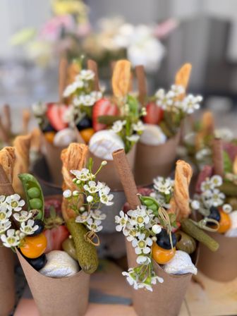 Kraft paper charcuterie cones: bite-sized grazing appetizers with cornichons, cherry tomatoes, black olives, mini round cheeses, pea pods, puff-pastry twists and breadsticks, garnished with small white flowers