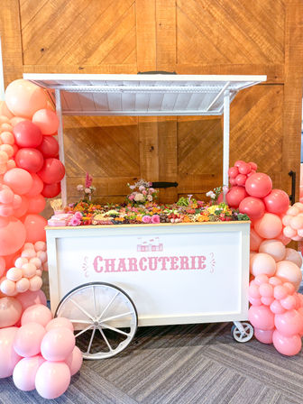 White charcuterie cart on wheels with colorful grazing board of fruits, cheeses and florals, framed by pink and coral balloon garlands against a rustic wooden wall.