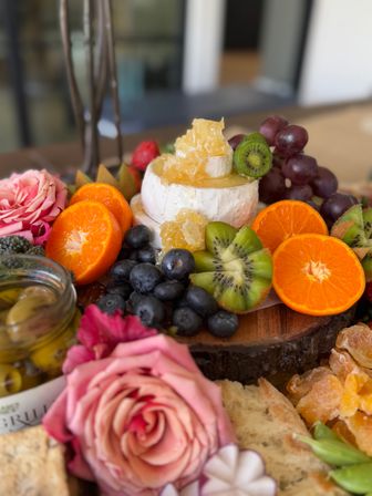 Bright charcuterie board on a wooden slab with a wheel of brie topped with honeycomb, sliced mandarins and kiwi, blueberries, grapes, olives and pink roses