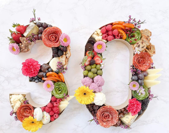 Playful gourmet charcuterie display shaped as the number 90, featuring salami roses, assorted cheeses, grapes, berries, olives, crackers, colorful vegetables and edible flowers on a light marble background.
