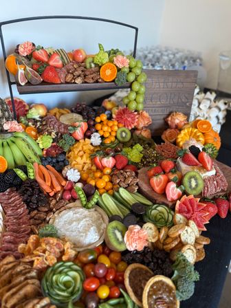 Vibrant charcuterie grazing board for events with assorted fruits (strawberries, kiwi, grapes, citrus), cheeses, cured meats, nuts, crackers and dip on a two-tier wooden display garnished with edible flowers.