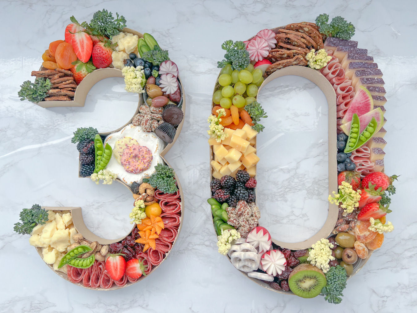 Number 30-shaped charcuterie grazing board on white marble, filled with assorted cheeses, sliced salami, grapes, strawberries, blackberries, kiwi, radishes, snap peas, olives, crackers, nuts, chocolate treats and a creamy dip — colorful party platter.