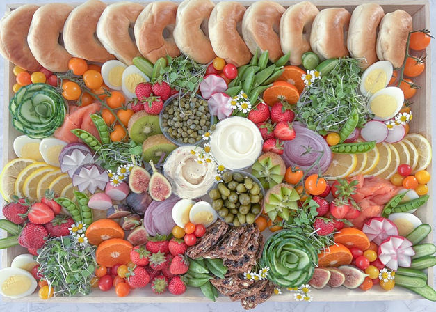 Vibrant bagel brunch board on a wooden tray: row of plain bagels, cream cheese and dips, smoked salmon, sliced hard-boiled eggs, cucumber rosettes, strawberries, figs, kiwi, lemon and orange slices, radishes, cherry tomatoes, snap peas, capers and seeded crackers in a colorful charcuterie-style spread