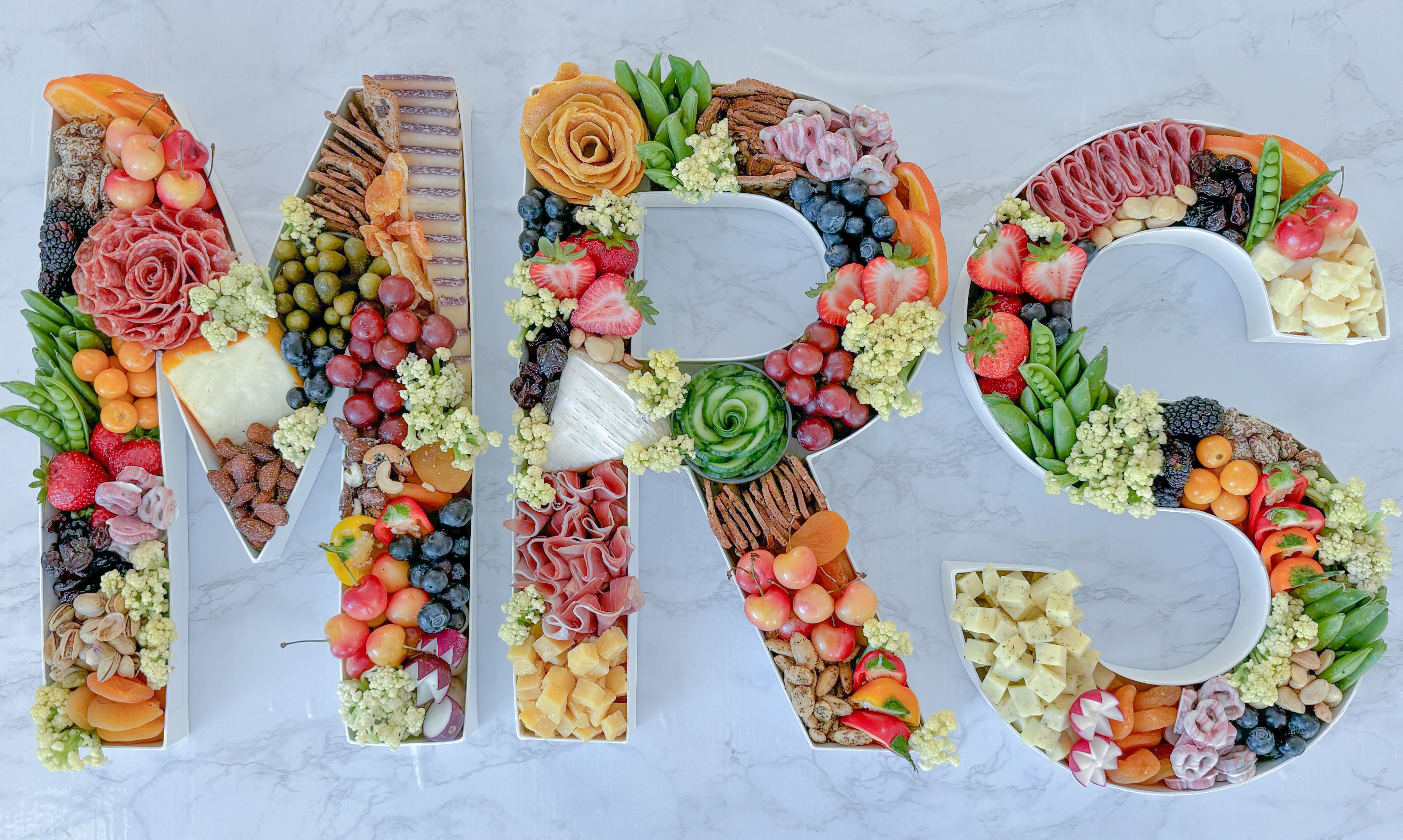 Letter-shaped 'MRS' charcuterie trays on marble filled with cheeses, cured meats, grapes, berries, nuts, crackers and edible flowers — playful wedding appetizer display.