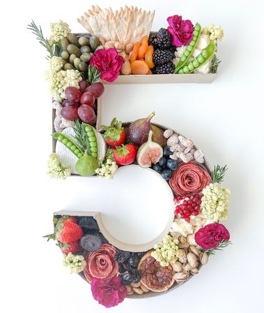 Number 5-shaped charcuterie display with crackers, salami roses, figs, strawberries, blackberries, grapes, dried apricots, snap peas, cheeses, nuts and edible flowers on a white background