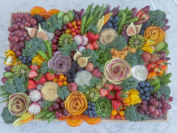 Vibrant rectangular fruit and vegetable grazing board on a wooden tray featuring carved vegetable roses, broccoli clusters, grapes, strawberries, blueberries, figs, kiwi, cherry tomatoes, snap peas, colorful mini peppers, orange slices and assorted dips.