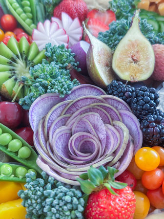 Close-up of a colorful produce platter with a purple radish arranged like a rose surrounded by figs, blackberries, strawberries, sliced kiwi, broccoli florets, sugar snap peas, cherries and yellow cherry tomatoes.