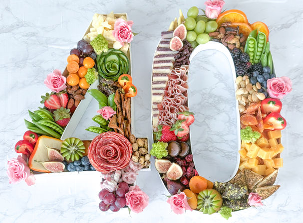 40-shaped charcuterie grazing board with a salami rosette, assorted cheeses, figs, grapes, berries, nuts, crackers, fresh vegetables and pink edible roses on a marble backdrop