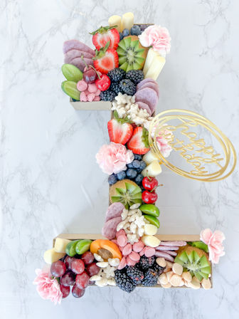 Number-one shaped birthday charcuterie board with strawberries, blackberries, kiwi, cherries, grapes, apricot, pastel candies, mini marshmallows and pink edible flowers on a white marble background