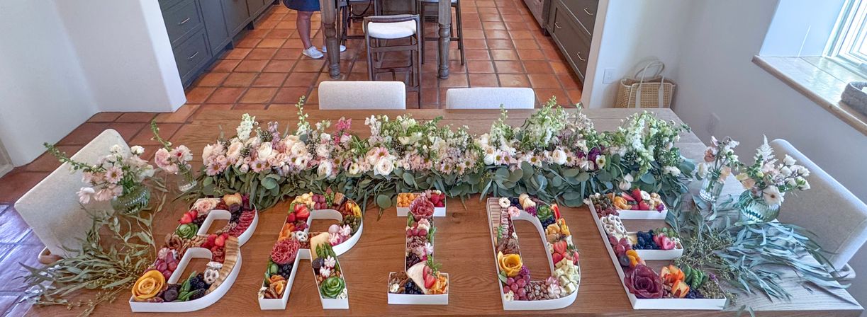 Charcuterie trays shaped as letters spelling BRIDE, filled with fruit, cheese, nuts and edible flowers on a wooden dining table, edged by a lush floral garland and greenery in a sunlit kitchen with terracotta tile floor — bridal shower display.