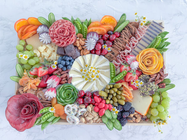 Vibrant artisan charcuterie and cheese board on marble: a brie wheel topped with chamomile, cured-meat roses, grapes, cherries, berries, olives, nuts, crackers, dried fruit and fresh veggies.