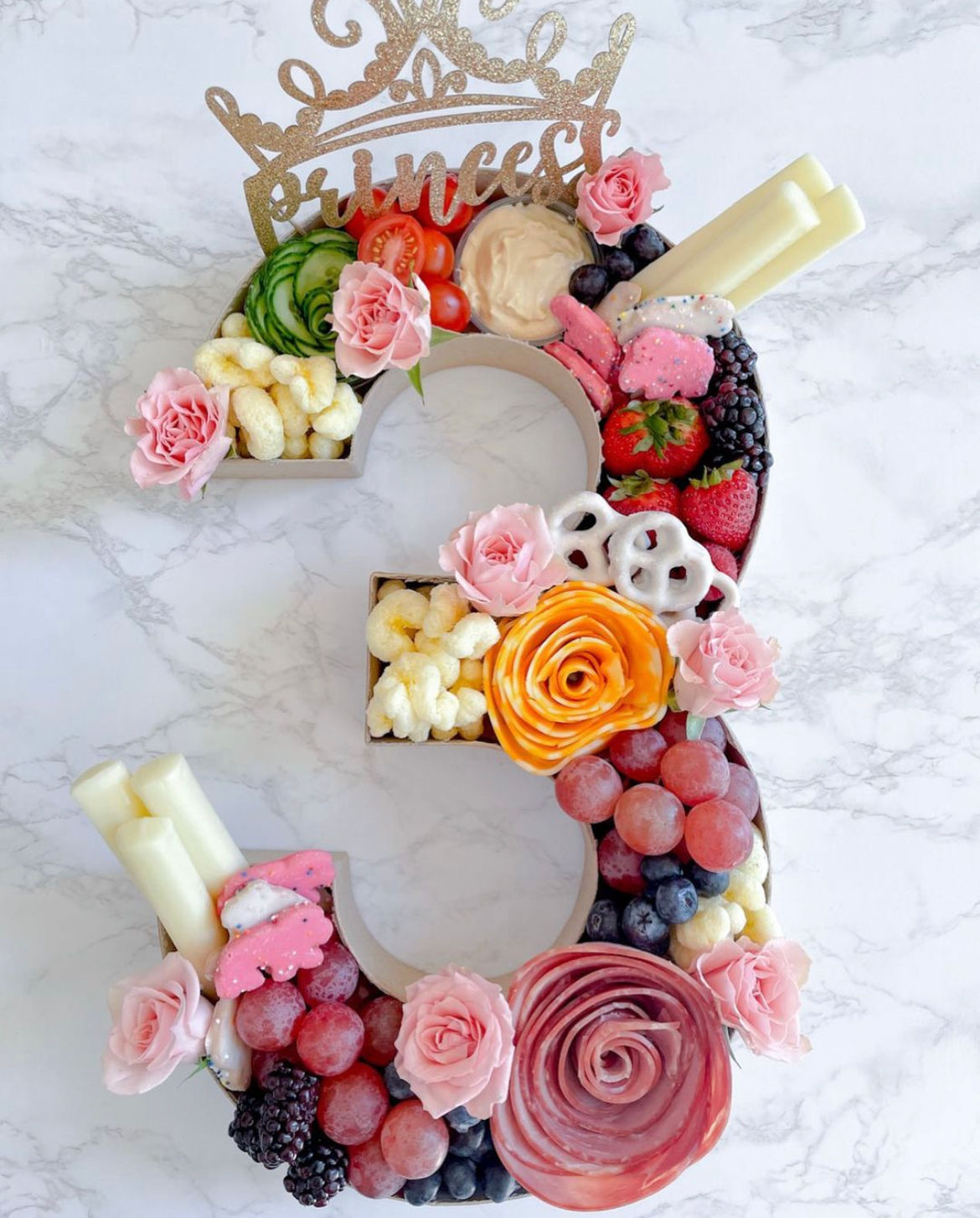 Princess-themed number 3 charcuterie board centerpiece with roses and rose-shaped salami, strawberries, grapes, blackberries, blueberries, cucumber roses, cherry tomatoes, cheese sticks, dips and yogurt-covered pretzels on marble