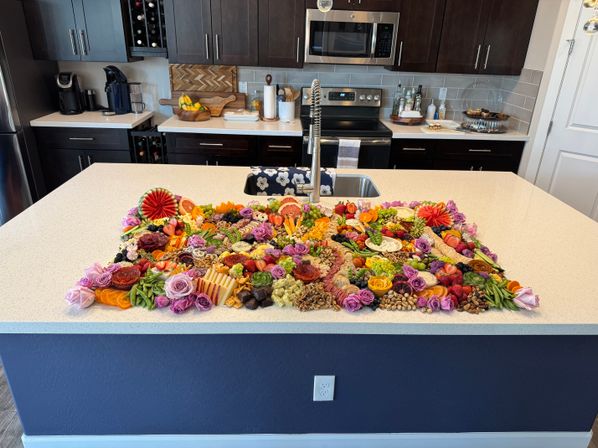 Vibrant oversized grazing charcuterie spread of cheeses, cured meats, fruits, nuts, crackers and edible flowers displayed on a white kitchen island in a modern home kitchen with dark cabinets and stainless appliances.