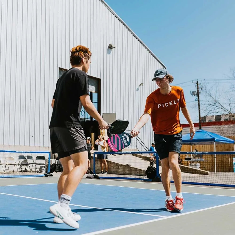 Two young players tapping paddles during an outdoor pickleball match on a blue court, sunny urban setting with a metal building and tents in the background.