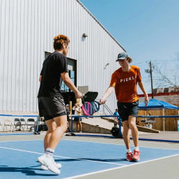 Two young players tapping paddles during an outdoor pickleball match on a blue court, sunny urban setting with a metal building and tents in the background.
