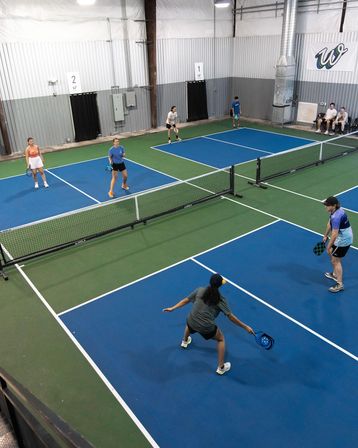 Overhead view of an indoor pickleball facility with multiple blue-and-green courts, players with paddles positioned for a doubles rally at the net in a recreational sports center.