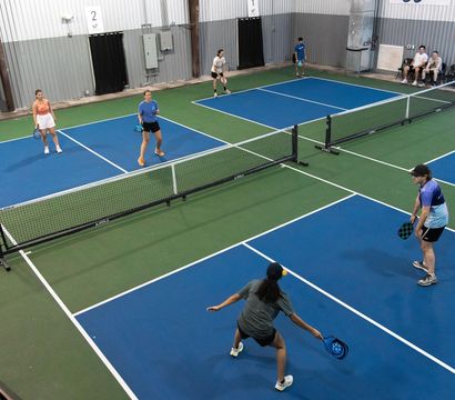 Overhead view of an indoor pickleball facility with multiple blue-and-green courts, players with paddles positioned for a doubles rally at the net in a recreational sports center.