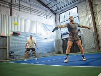 Two players on an indoor pickleball court — one lunging to hit a yellow ball with a paddle on a blue-and-green court inside a metal-paneled gym with large windows