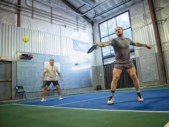 Two players on an indoor pickleball court — one lunging to hit a yellow ball with a paddle on a blue-and-green court inside a metal-paneled gym with large windows
