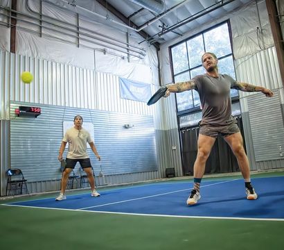 Two players on an indoor pickleball court — one lunging to hit a yellow ball with a paddle on a blue-and-green court inside a metal-paneled gym with large windows