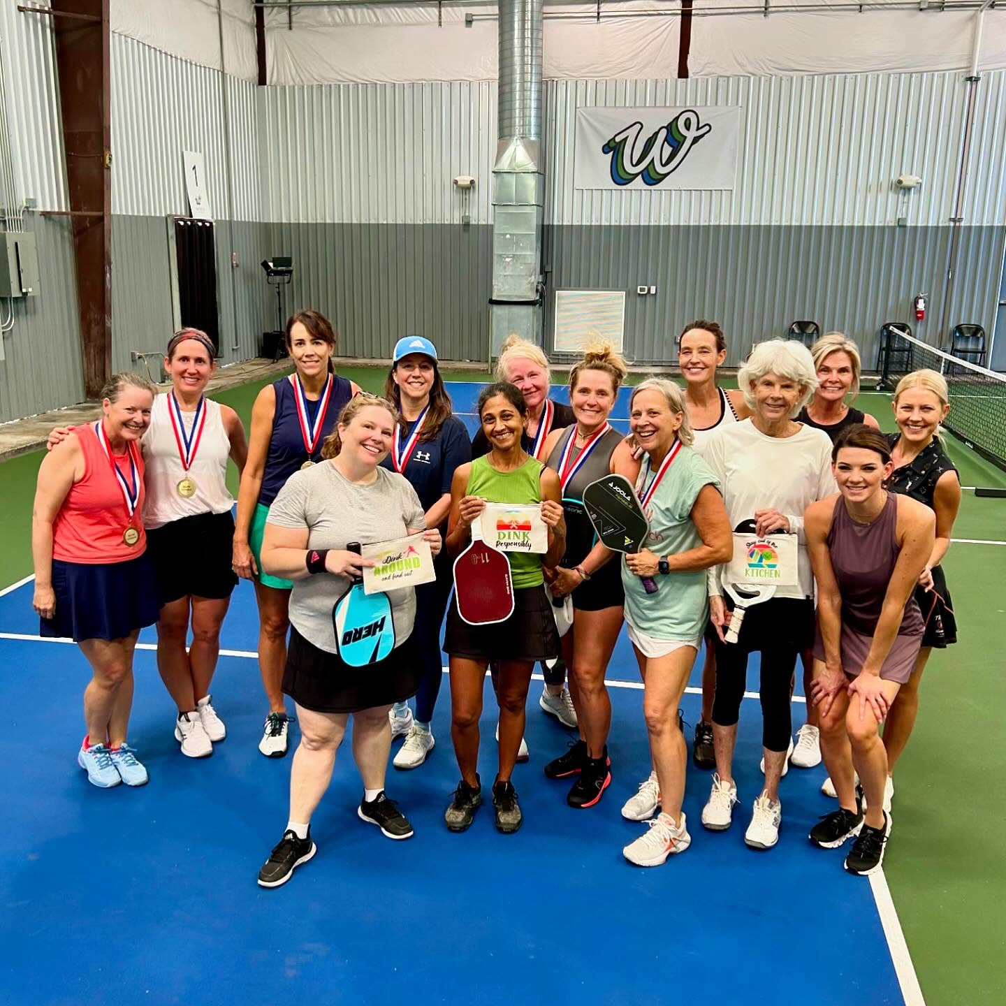 Smiling women’s pickleball group on an indoor court celebrating a tournament, holding paddles, medals and award signs — recreational players posing after a match