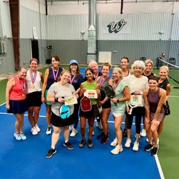 Smiling women’s pickleball group on an indoor court celebrating a tournament, holding paddles, medals and award signs — recreational players posing after a match