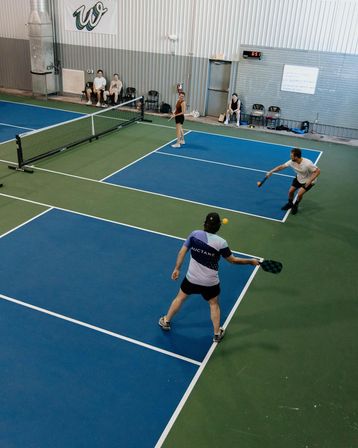 Four players in a doubles pickleball match on a blue-and-green indoor court at a recreational sports facility, two near the net and two returning shots with paddles and an orange ball, with spectators seated along the wall.